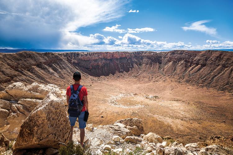 A hiker in a backpack and red shirt stands on the rim of the 50,000 year-old Meteor Crater