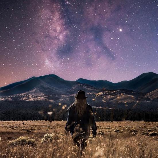 A woman stands in a field with her back to the camera looking out at mountains in Flagstaff under the Milky Way 